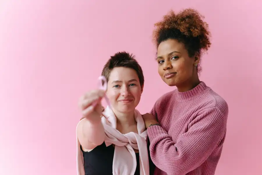 Duas mulheres diante de uma parede rosa; uma delas segura a fita rosa do câncer de mama