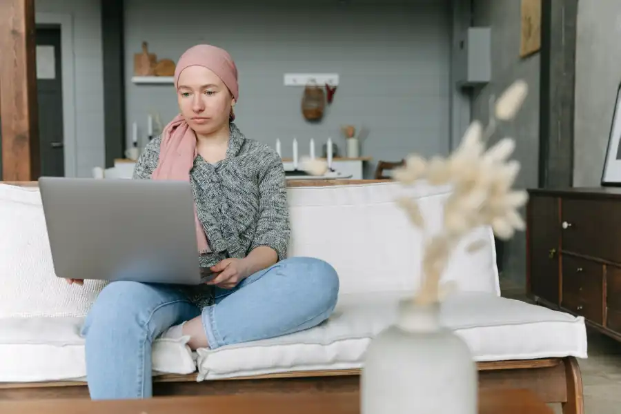 Mulher de lenço rosa trabalhando no notebook, sentada num sofá claro de uma sala aconchegante