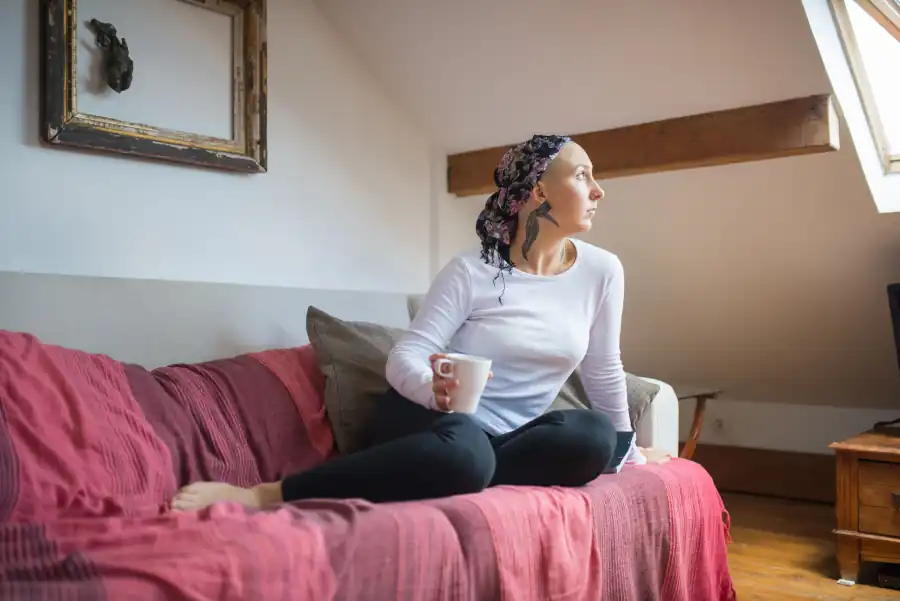Mulher de lenço sentada num sofá vermelho, segurando uma caneca e olhando pela janela