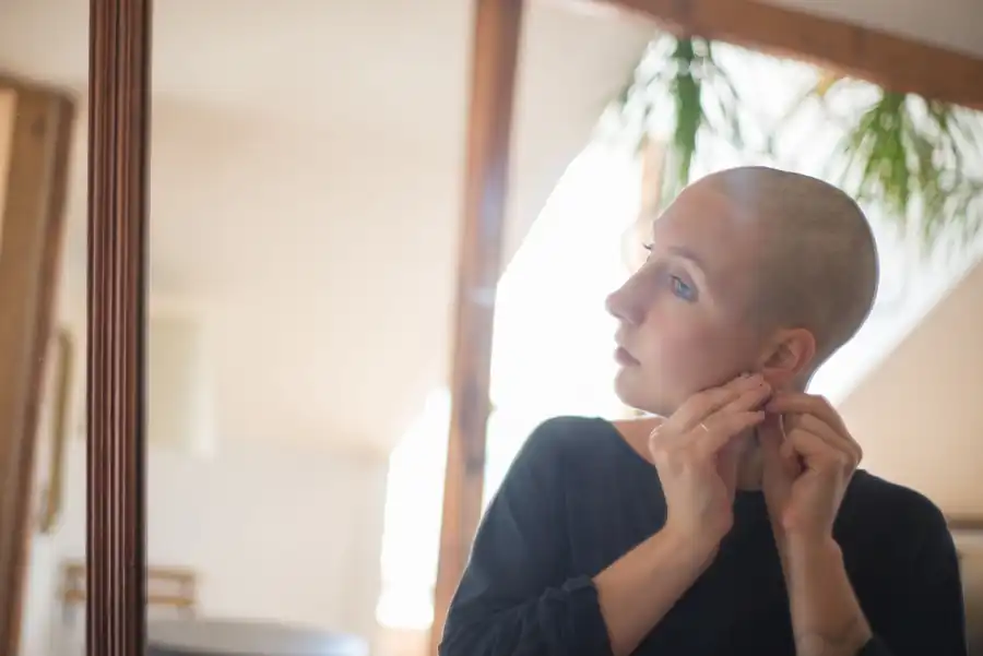 Mulher careca colocando um brinco, perto de uma janela iluminada